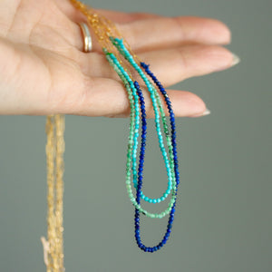 Hand holding three beaded necklaces in green, blue, and gold against a neutral background